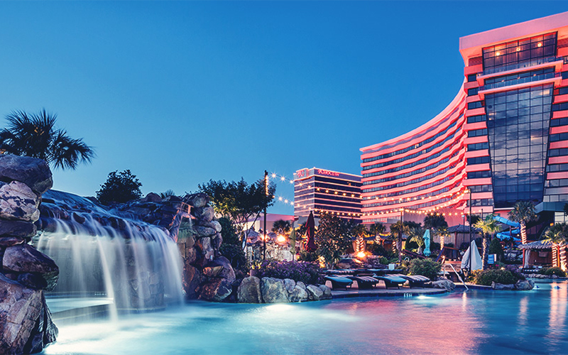 The outside of a casino with big glass windows and red lights with a waterfall, reflecting pool, and palm trees outside. 