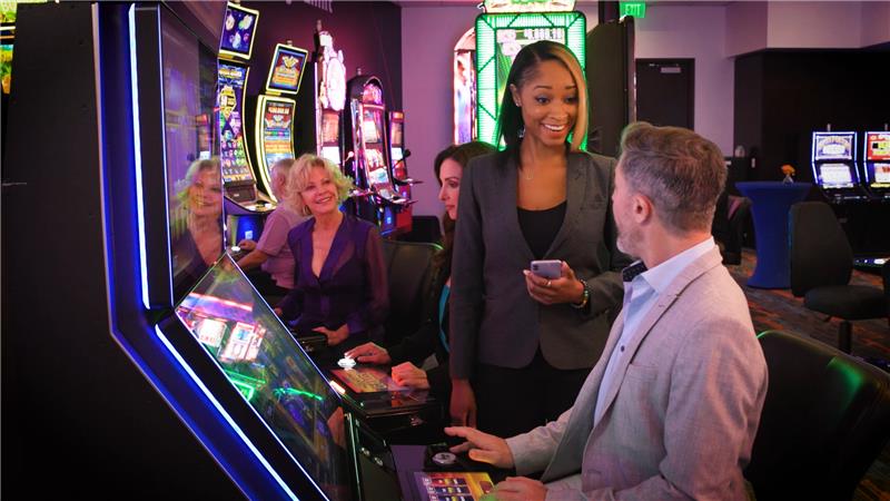 A woman in a gray blazer talking to a man with a gray beard sitting at an IGT slot machine in a crowded room full of IGT's gaming machines and titles.