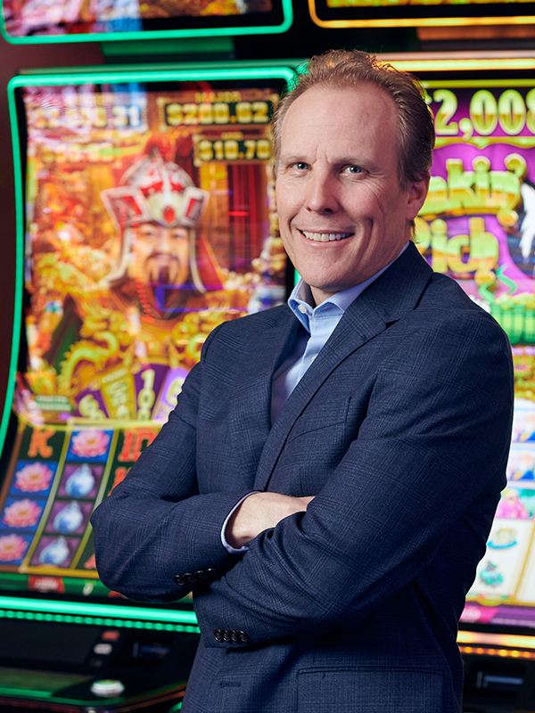 Photo of Roger similing with his arms crossed in front of two video slot cabinets wearing a navy suit jacket and blue collared shirt.