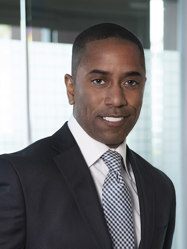 Photo of Darnell smiling in front of a glass window wearing a black suit jacket, white collared shirt, and black and white tie. 